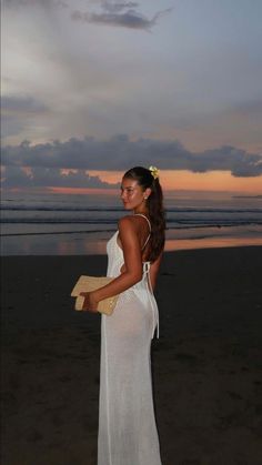 a woman in a white dress standing on the beach at sunset with her back to the camera