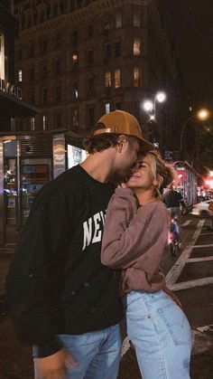 a man and woman standing next to each other in the street at night with buildings behind them