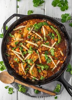 a skillet filled with meat and vegetables on top of a white wooden table next to spoons