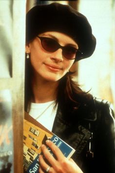 a woman wearing sunglasses and a hat holding a book in her hands while standing next to a pole