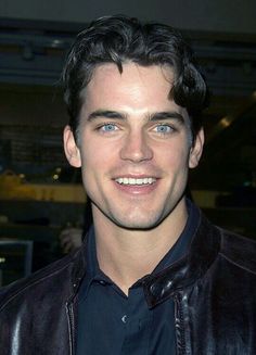 a close up of a person wearing a black shirt and leather jacket smiling at the camera