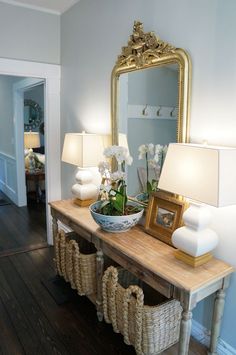 a wooden table topped with a bowl of flowers next to a mirror and lamp on top of a hard wood floor