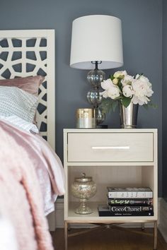 a nightstand with flowers and books on it next to a white bed in a bedroom