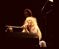 a woman sitting at a piano in front of a man with long hair on stage