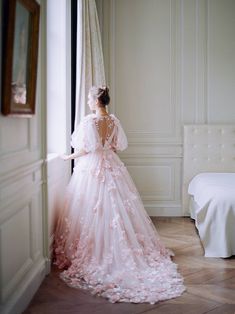 a woman in a pink wedding dress standing by a window with her back to the camera