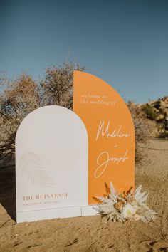 an orange and white wedding sign in the desert with flowers on it's side