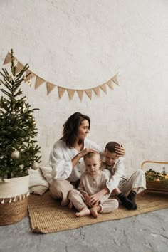 a woman and two children sitting on the floor in front of a christmas tree with decorations