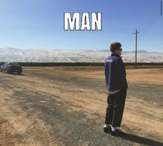 a man standing on the side of a dirt road next to a blue car and mountains