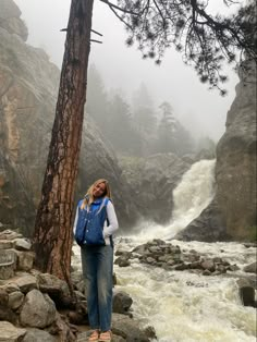 a woman standing next to a tree on top of a rock covered hillside near a river