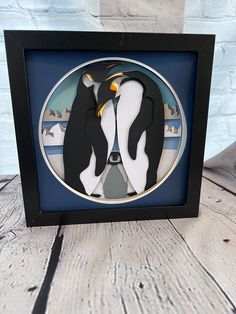 two penguins standing next to each other in a black frame on a wooden table with white brick wall behind them