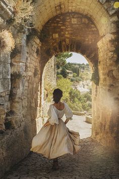 a woman in a white dress is walking through an old stone tunnel with sunlight streaming through it