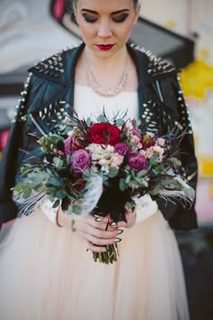 a woman in a white dress and black leather jacket holding a bouquet with red flowers