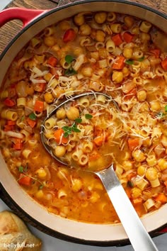 a pot filled with pasta and vegetables on top of a wooden table next to bread