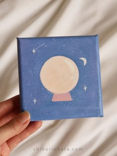 a hand is holding up a small painting on a blue background with stars and the moon