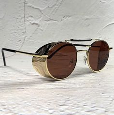 These Brown 1910s Steampunk Sunglasses feature a gold metal frame with side shields, blending avant-garde Victorian and goth styles. Perfect for a cyberpunk aesthetic, these industrial shades make a unique gift idea. New old stock. Great condition. Never worn. UV Protection. NOS Deadstock Made in Europe Free combined shipping worldwide. FIT INFORMATION 50 mm Eye Size 22 mm Bridge 145 mm Temple/Arm Width 5'' 1/4 Inches Height 1'' 7/8 Inches Vintage Sunglasses With Metal Frame For Outdoor, Retro Gold Sunglasses With Mirrored Lenses, Gold Retro Sunglasses With Mirrored Lenses, Steampunk Sunglasses With Mirrored Lenses, Retro Gold Shield Sunglasses With Mirrored Lenses, Retro Gold Shield Sunglasses With Tinted Lenses, Gold Anti-reflective Sunglasses For Outdoors, Gold Shield Sunglasses With Tinted Lenses For Outdoor, Vintage Gold Sunglasses With Metal Frame