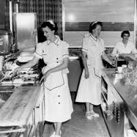 two women in white aprons are standing at the counter and one woman is wearing an apron