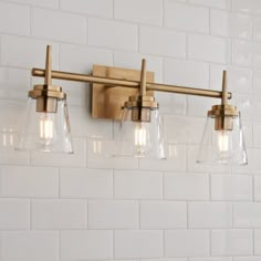 three light bathroom fixture with clear glass shades on white tile wall behind the lights is a brass finish