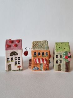 three small ceramic houses sitting next to each other on a white surface with an apple hanging from the roof