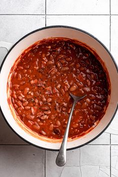 a pot filled with chili and beans on top of a white tile floor next to a spoon