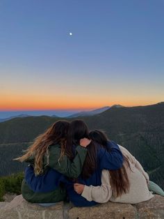 two women sitting on top of a rock with their arms wrapped around each other looking at the sunset