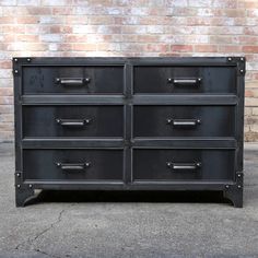 a black dresser with metal handles and drawers on the bottom, in front of a brick wall