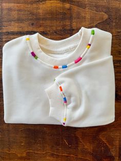 a white t - shirt with multicolored beads on the front and back, sitting on a wooden surface