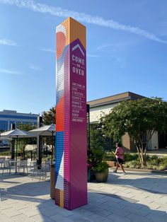 a tall colorful sign sitting on the side of a road next to a sidewalk with tables and umbrellas