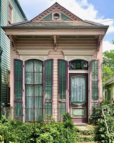 an old pink house with green shutters on the front and side windows, surrounded by greenery