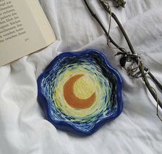 a blue bowl sitting on top of a bed next to an open book and twig