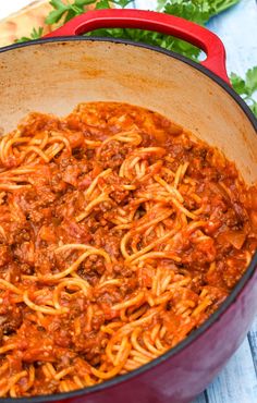 a red pot filled with spaghetti and sauce on top of a wooden table next to parsley