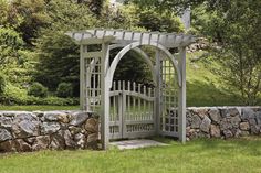 a stone wall with a white wooden arbor in the middle and green grass around it