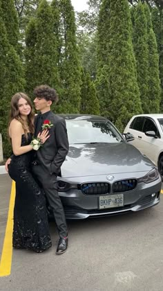 a man and woman standing in front of a silver car with trees behind them on the street