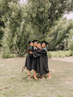 three women in graduation gowns hugging each other on the grass with trees in the background