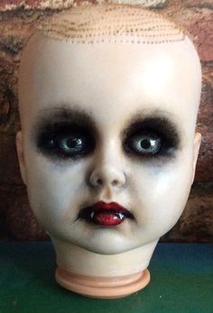 a creepy doll head with black eyes and red lips on a green table next to a brick wall