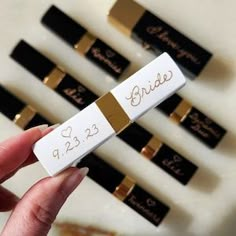 a person holding up some white and gold lipstick in front of several black and gold ones