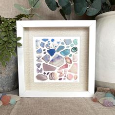 a white frame with some sea glass in it next to rocks and a potted plant