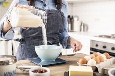 a woman pouring milk into a bowl on top of a counter next to eggs and other ingredients