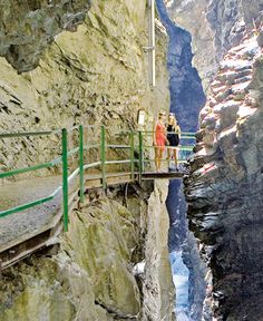 two people are standing on a bridge over a narrow canyon in the mountains above them