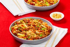 two bowls filled with noodles and vegetables on top of a red table cloth next to chopsticks