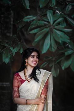 a woman wearing a white sari and gold jewelry standing in front of a tree