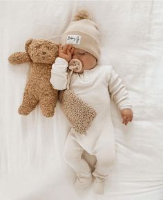 a baby laying on top of a bed next to a teddy bear wearing a hat