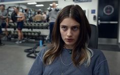 a woman sitting in a gym looking at the camera with other people standing behind her