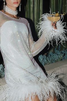 a woman in a white dress holding a wine glass with feathers around her neck and wearing pearls