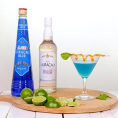 a wooden tray topped with drinks next to a bottle of blue liquid and limes