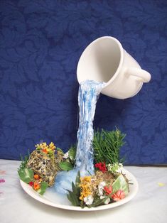 a white coffee cup pouring water into a plate with flowers and plants on it, in front of a blue wall