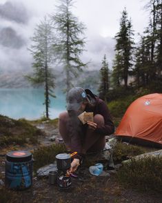 a man sitting on top of a field next to a tent and camping gear in front of a lake