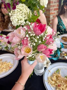 a woman holding a vase filled with flowers next to plates of food on a table