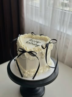 a white cake with black ribbon on top sitting on a table in front of a window