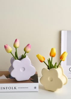 two vases with yellow and pink flowers in them next to a book on a table