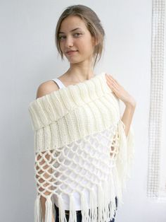 a woman standing in front of a white wall wearing a knitted shawl with fringes